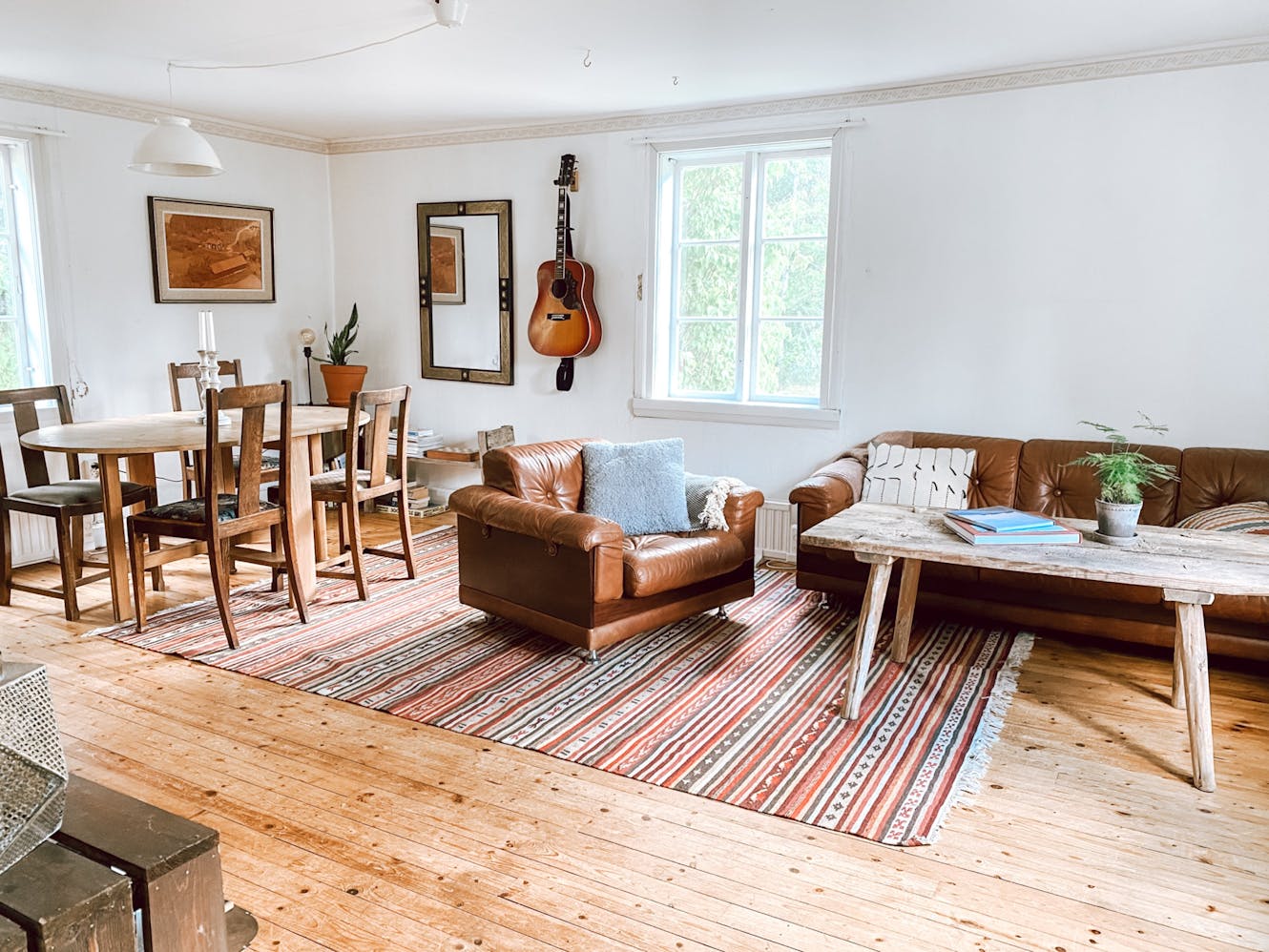 Living room and dining area with brown leather sofa, wooden dining table with chairs, rustic coffee table, striped area rug, and guitar mounted on white wall