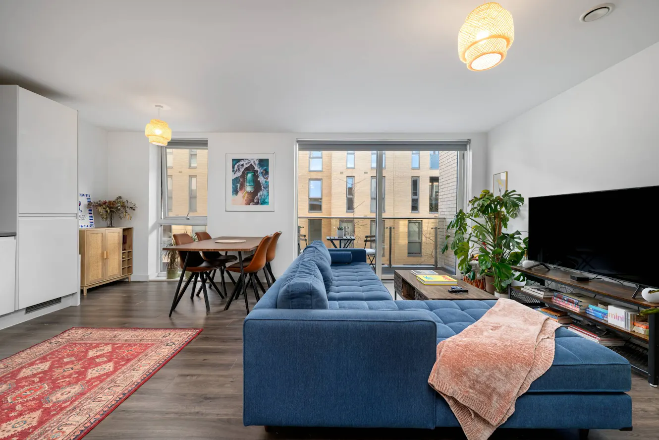 Living room with blue sectional sofa, dining table with orange chairs, large windows with balcony access, TV console, and hardwood floors