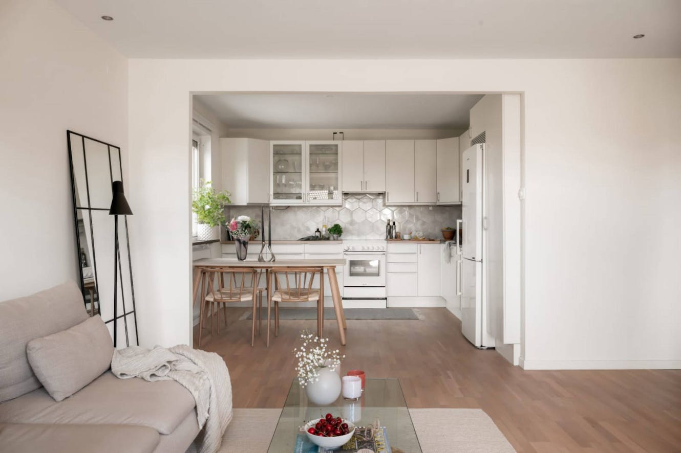 Living room with beige sofa and coffee table, view through to kitchen with white cabinets, dining table with wooden chairs, and hexagonal tile backsplash