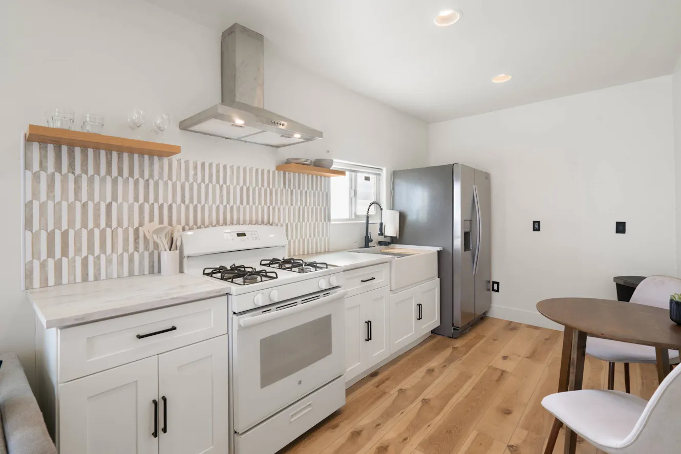 Kitchen with white cabinets, gas range, stainless steel refrigerator, geometric tile backsplash, wooden shelving, and light wood flooring