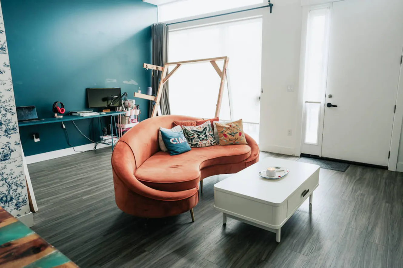 Living room with coral curved sofa, white coffee table, desk with monitor and headphones, wooden clothing rack, and entry door with natural light