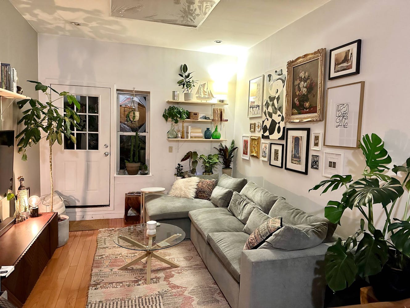 Living room with sage green sectional sofa, glass coffee table, gallery wall with framed artwork, floating shelves, indoor plants, and hardwood floors