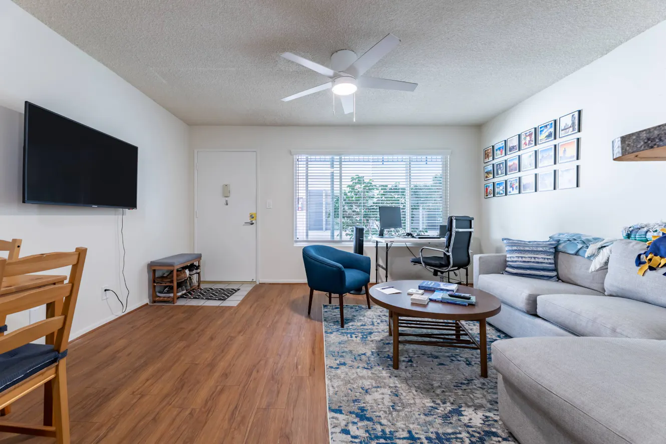 Living room with gray sofa, blue accent chair, wooden coffee table, wall-mounted TV, workspace desk with office chair, and ceiling fan