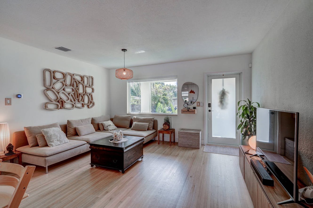 Living room with beige sectional sofa, wooden coffee table, light wood flooring, large window with natural light, wall-mounted TV, and glass entry door