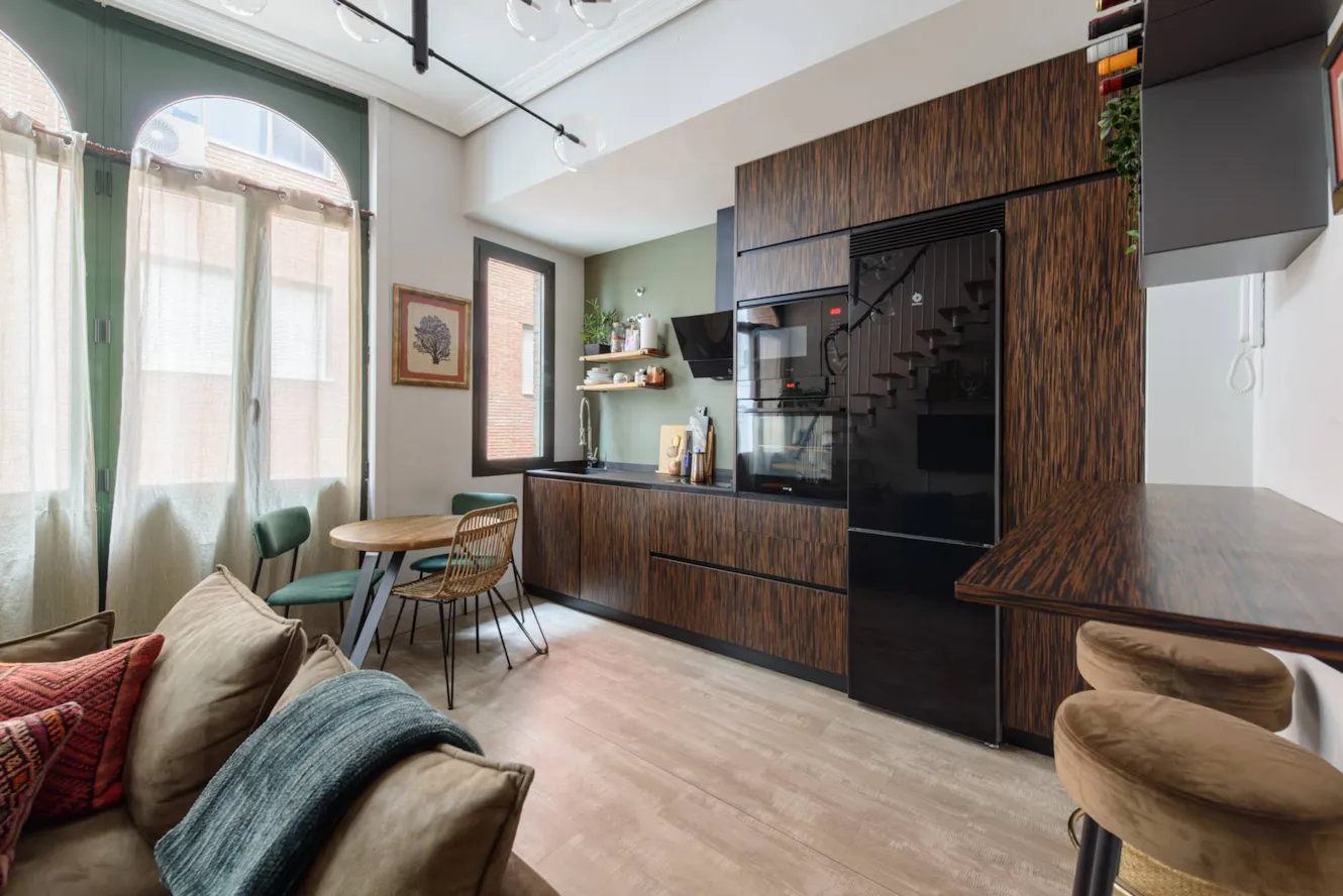 Open-concept living space with kitchen featuring wood-grain cabinets, black appliances, dining table with green chairs, sofa, and large arched window with natural light