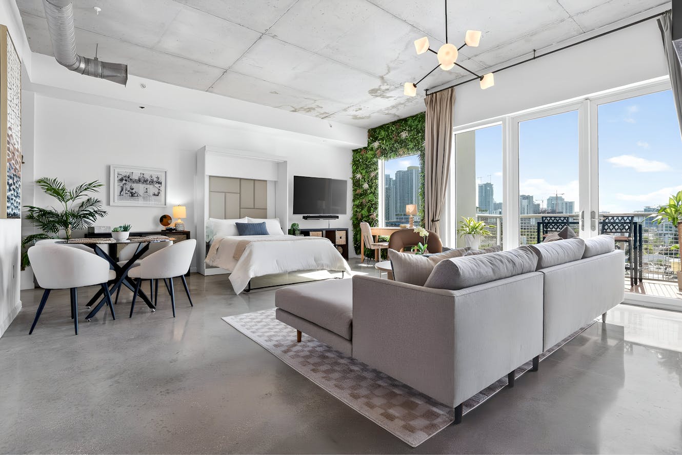 Modern loft studio with gray sectional sofa, dining table, queen bed, wall-mounted TV, living wall, floor-to-ceiling windows with city views, and exposed concrete ceiling