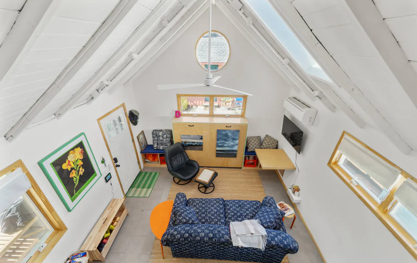 Living room with vaulted white ceiling, skylights, blue patterned sofa, black rocking chair, wooden storage cabinets, wall-mounted TV, and round mirror window