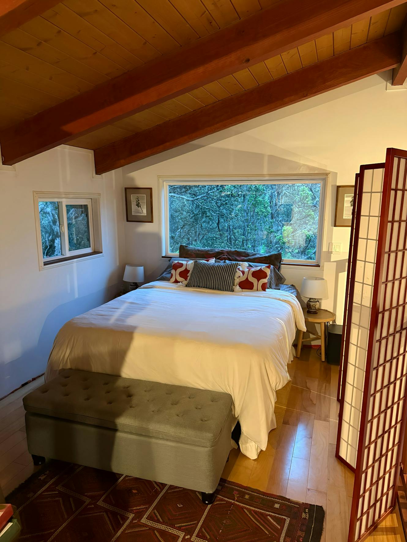 Bedroom with queen bed, exposed wood beam ceiling, large window with tree views, nightstands with lamps, and upholstered bench at foot of bed