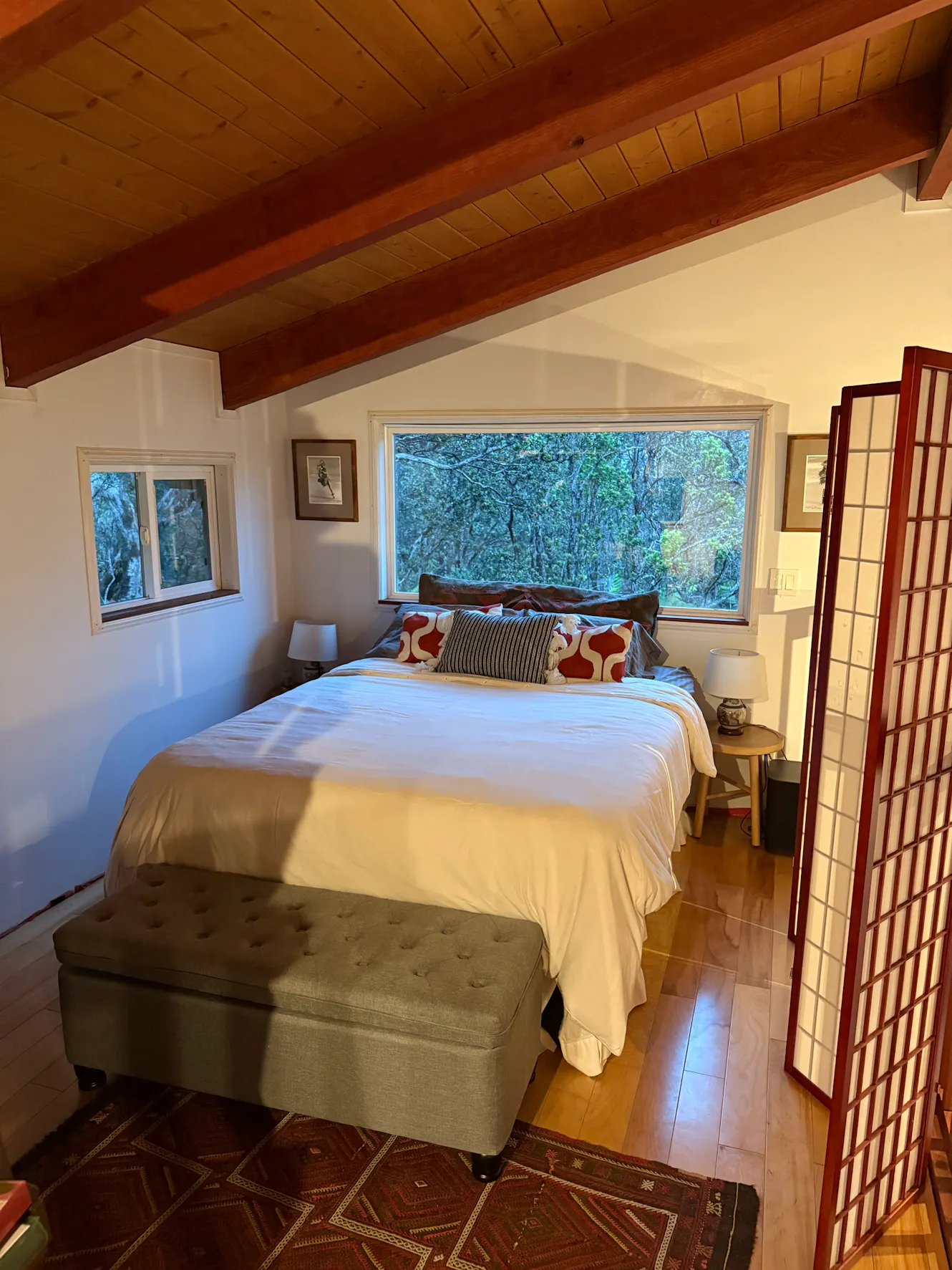 Bedroom with queen bed, exposed wood beam ceiling, large window with tree views, nightstands with lamps, and upholstered bench at foot of bed
