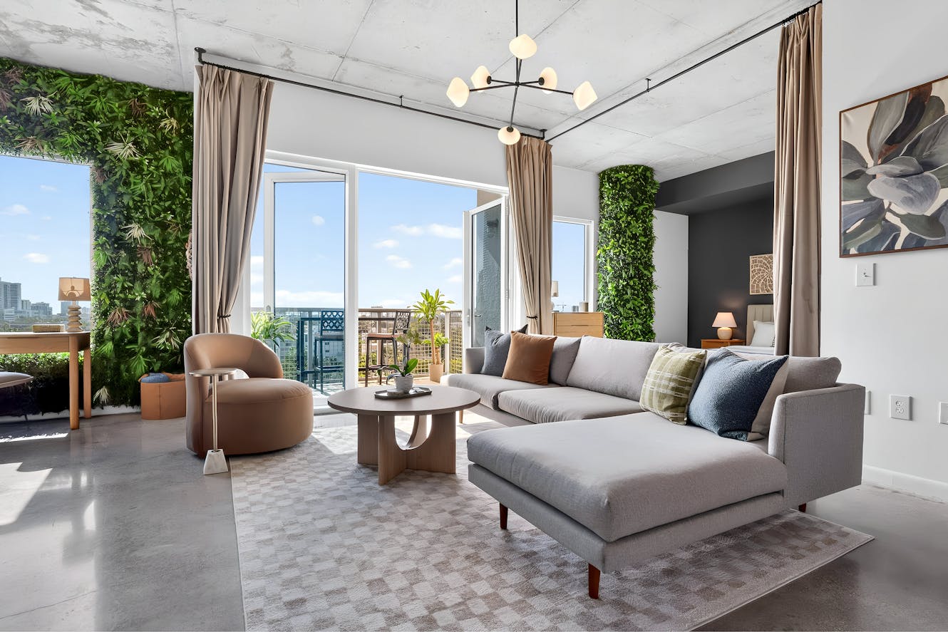Living room with gray sectional sofa, tan accent chair, round coffee table, floor-to-ceiling windows with city views, exposed concrete ceiling, and vertical garden walls