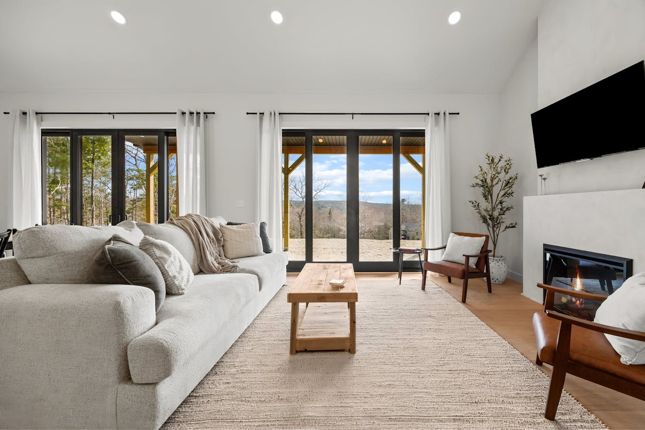 Living room with gray sectional sofa, wooden coffee table, accent chair, wall-mounted TV, modern fireplace, and floor-to-ceiling windows with mountain views