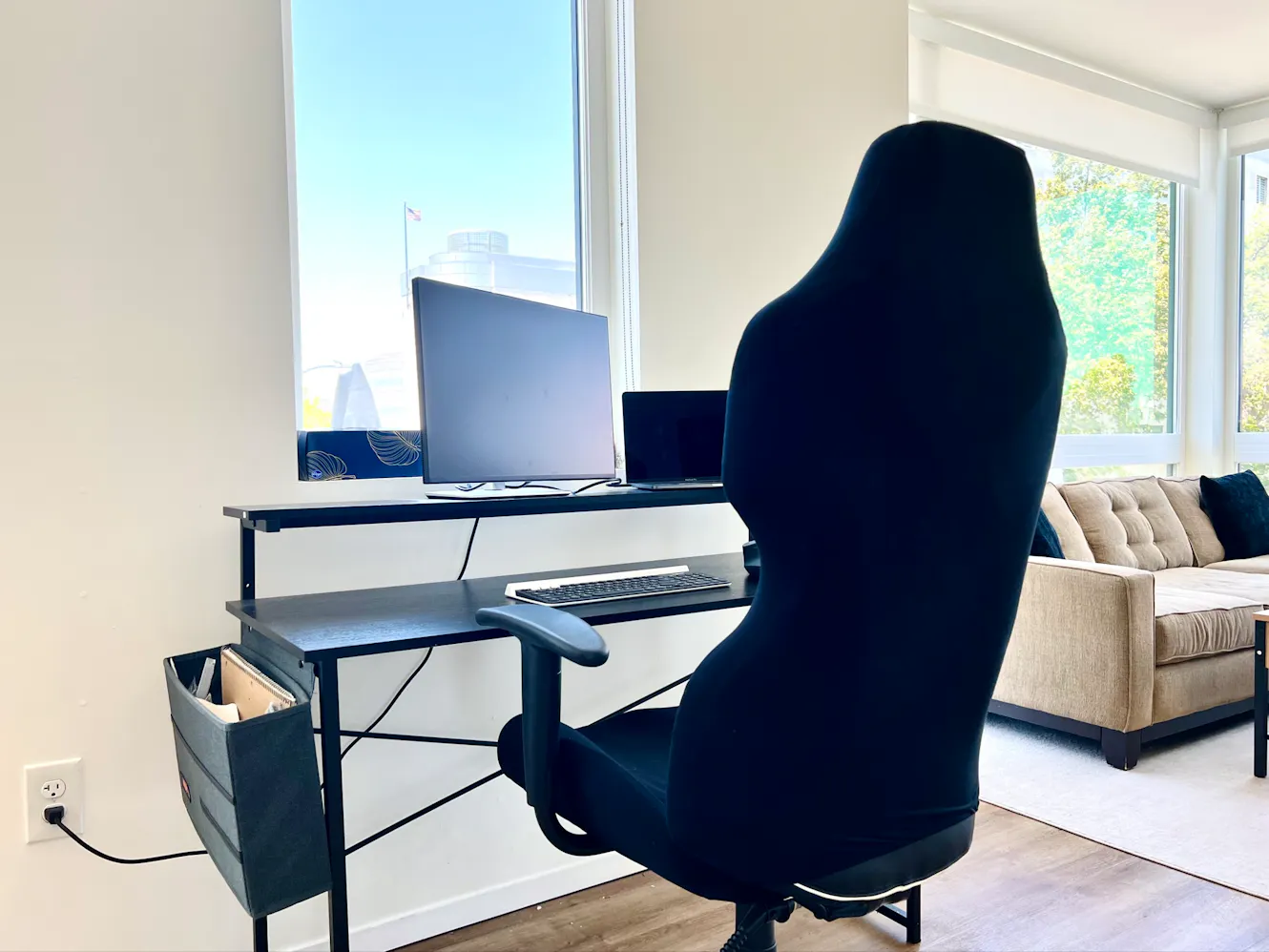 Living room workspace with black desk, laptop, ergonomic office chair, large windows with natural light, and beige sofa
