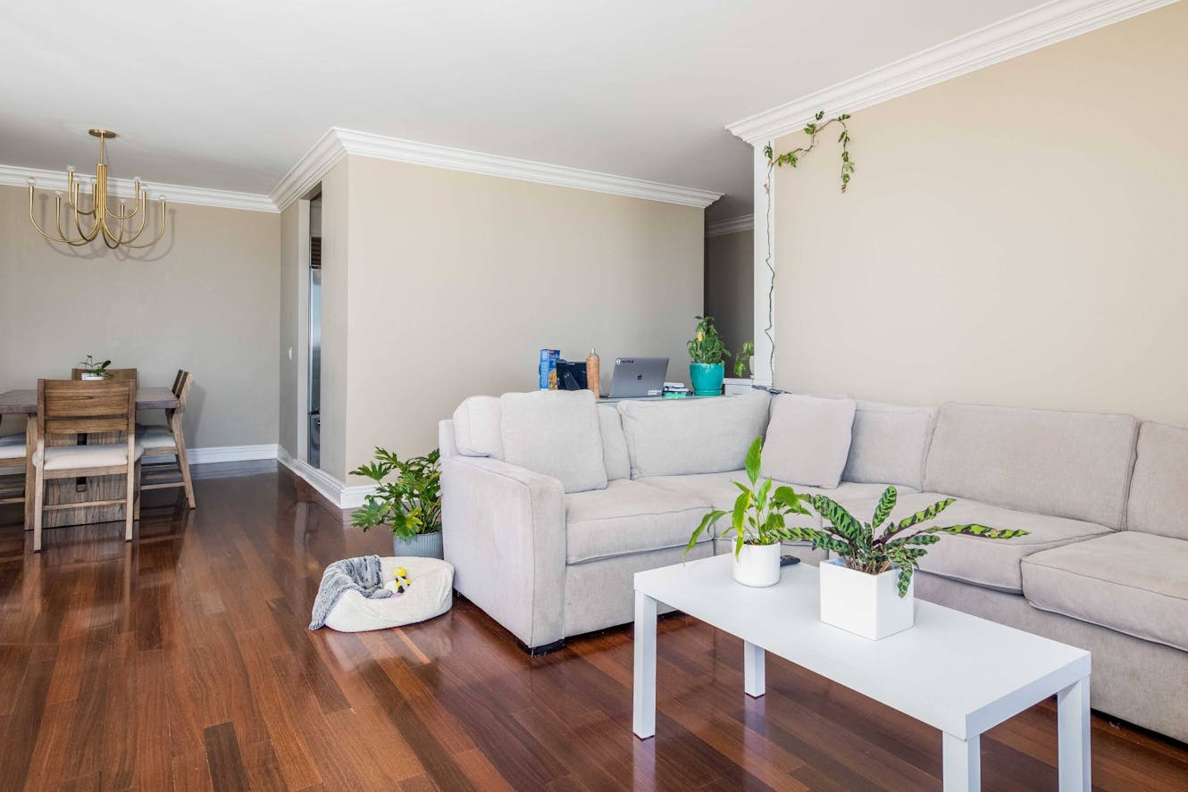 Living room with beige sectional sofa, white coffee table with plants, dark hardwood floors, and adjacent dining area with wooden table and chandelier