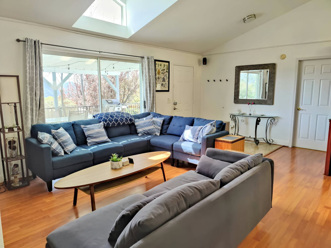 Living room with blue sectional sofa, gray sofa, wooden coffee table, large windows with skylight, and view of outdoor patio