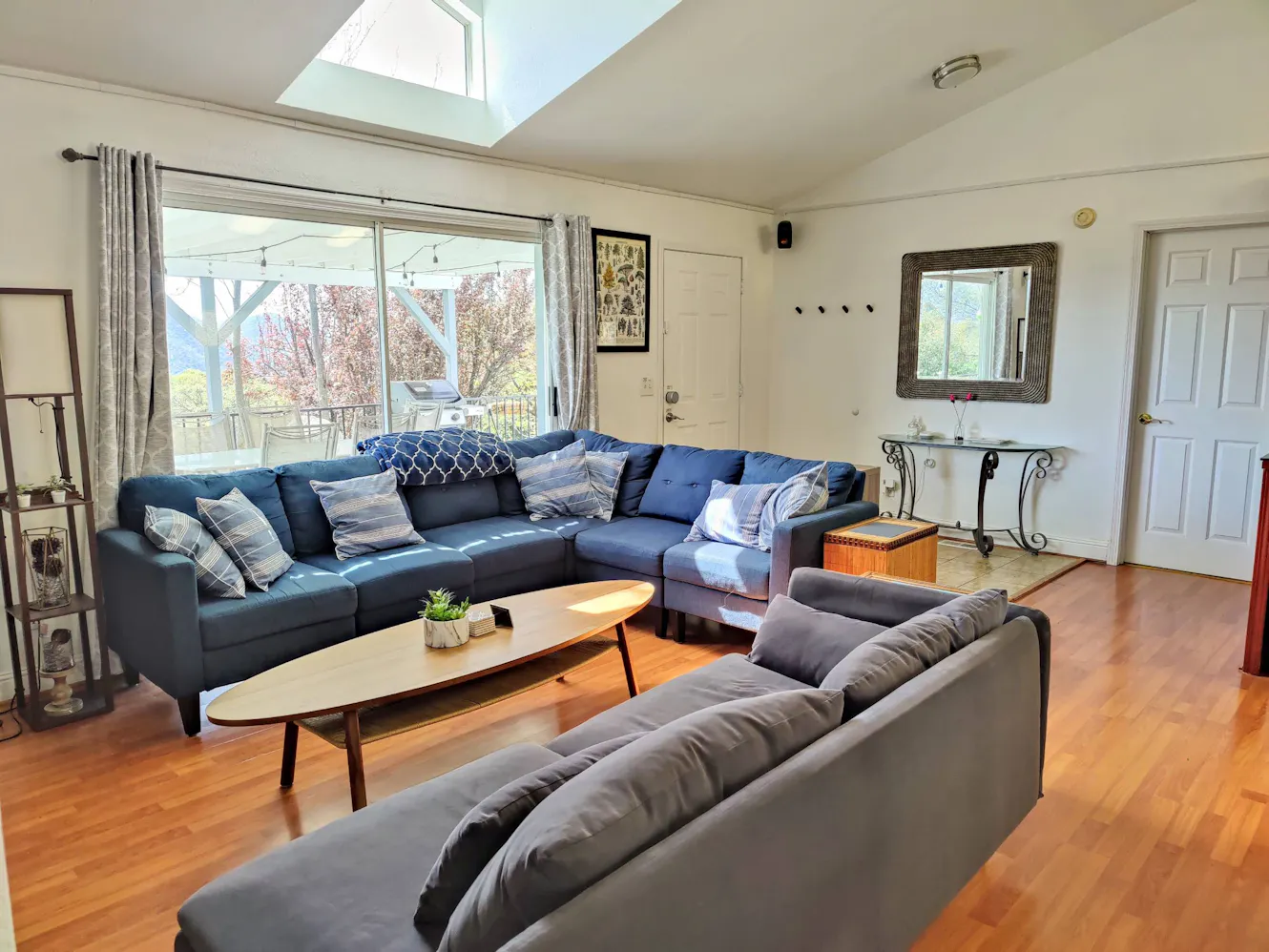 Living room with blue sectional sofa, gray sofa, wooden coffee table, large windows with skylight, and view of outdoor patio