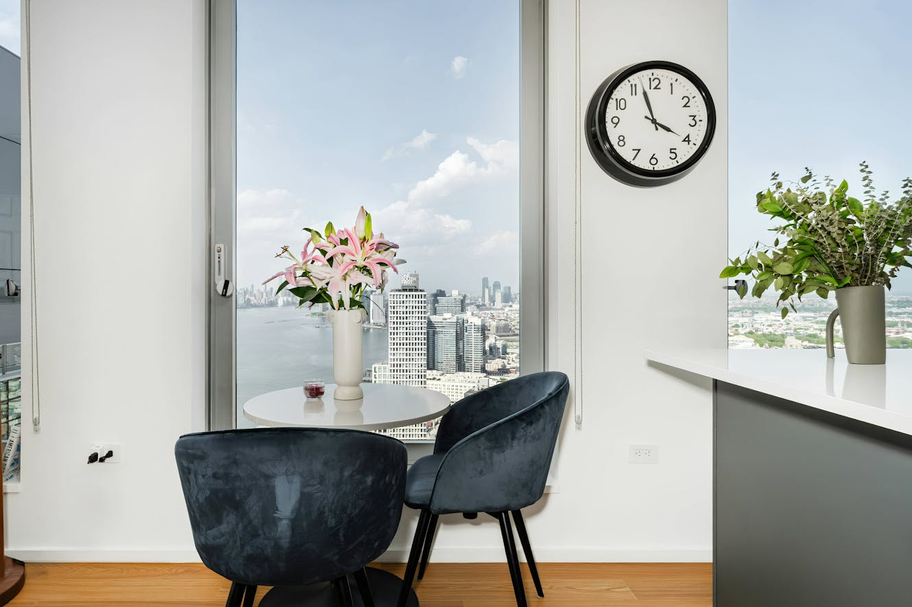 Dining area with round white table, two dark blue velvet chairs, floor-to-ceiling windows with city and water views, wall clock, and fresh flowers