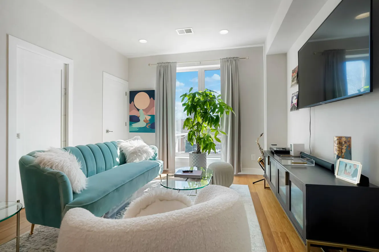 Living room with teal velvet sofa, white furry ottoman, glass coffee table, wall-mounted TV, large window with natural light, and potted plant