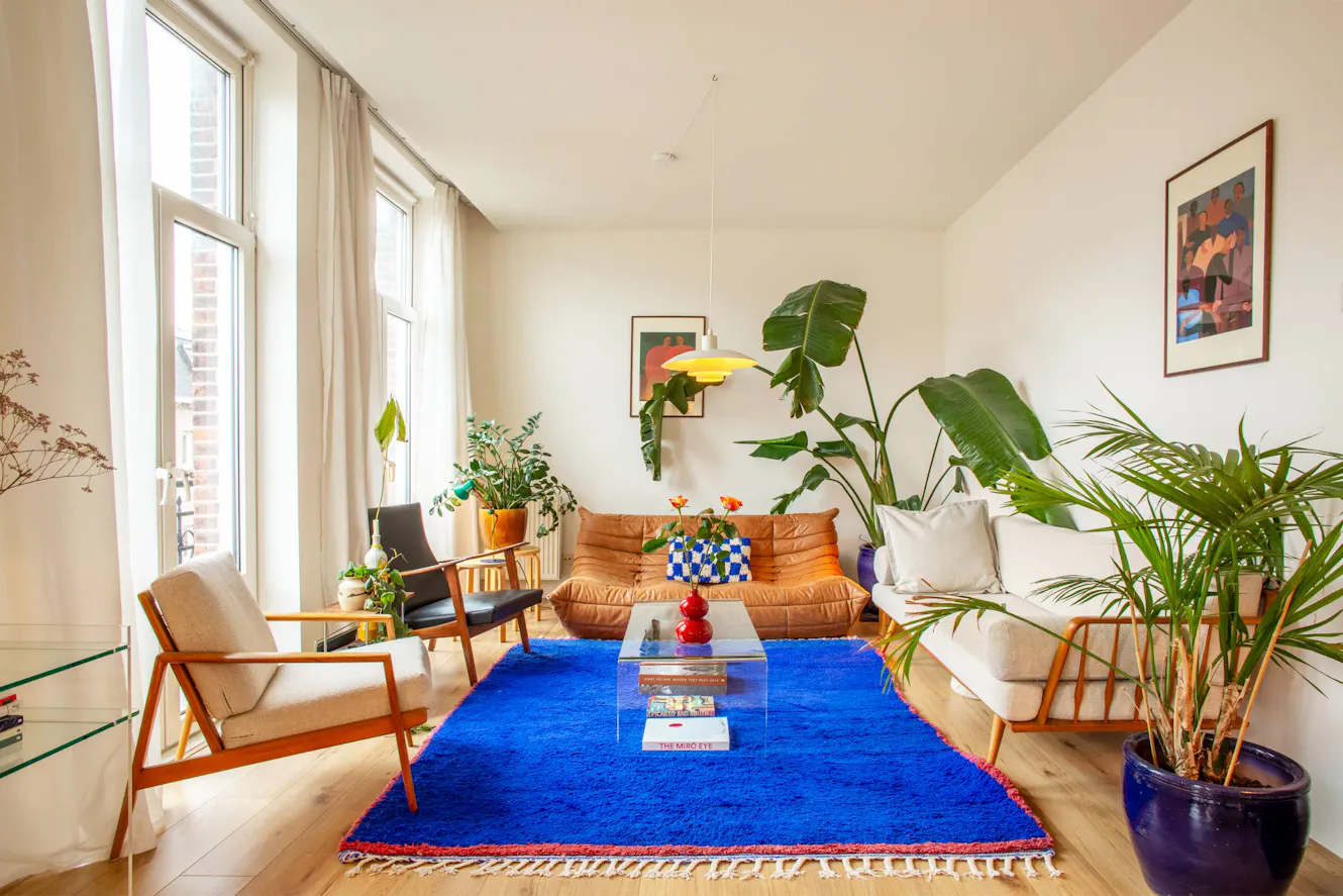 Living room with orange leather sofa, mid-century modern chairs, glass coffee table on bright blue rug, large windows with natural light, and tropical plants