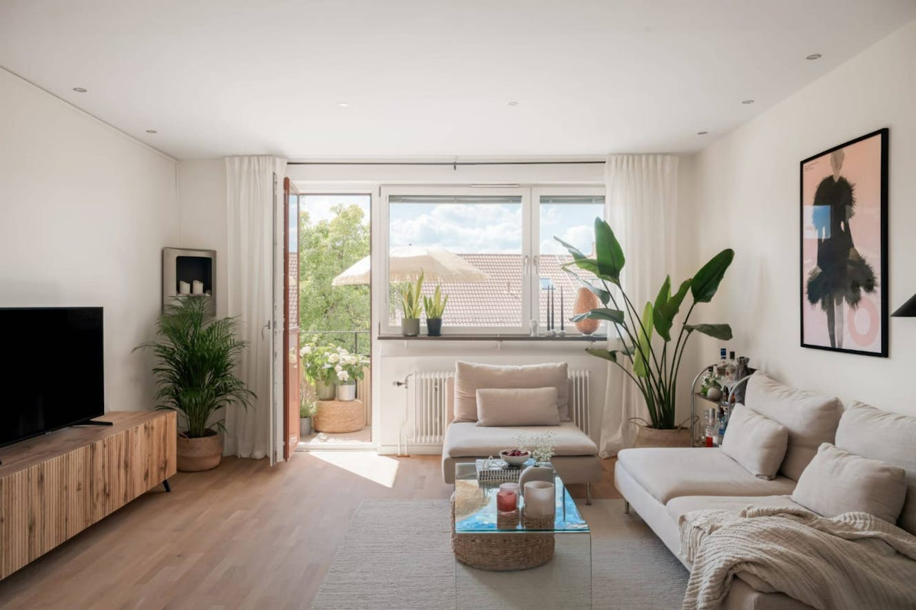 Living room with white sectional sofa, glass coffee table, large windows with natural light, indoor plants, wall-mounted TV, and wooden media console