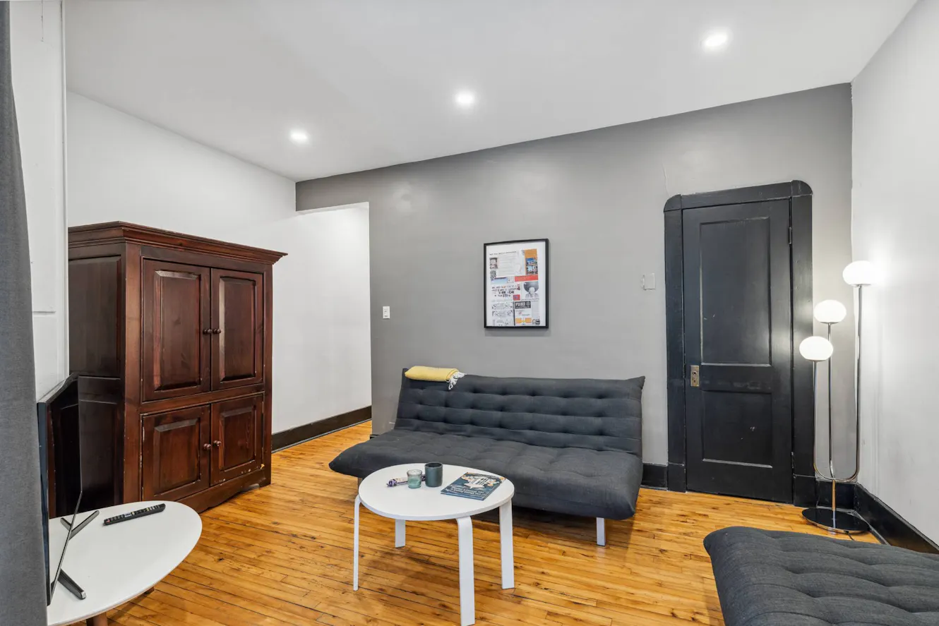 Living room with gray futon sofa, white round coffee table, hardwood floors, dark wood armoire, and modern floor lamp