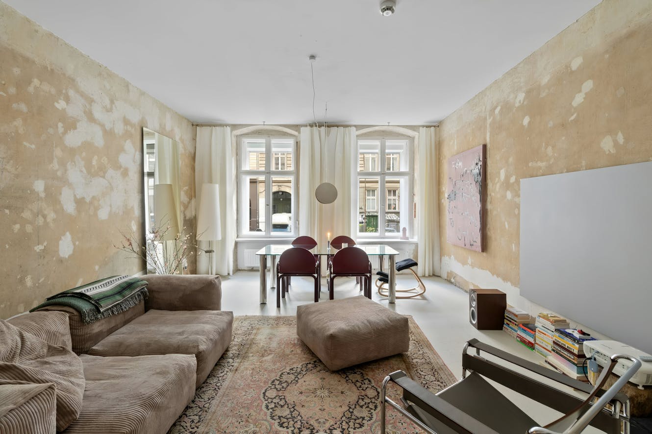 Living room with brown corduroy sofa, ottoman, glass dining table with burgundy chairs, arched windows with white curtains, and distressed tan walls