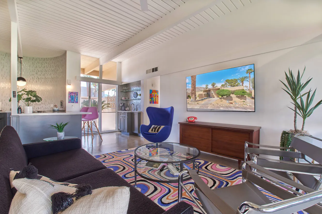 Living room with dark sectional sofa, blue accent chair, glass coffee table, wall-mounted TV, wooden credenza, and colorful patterned rug