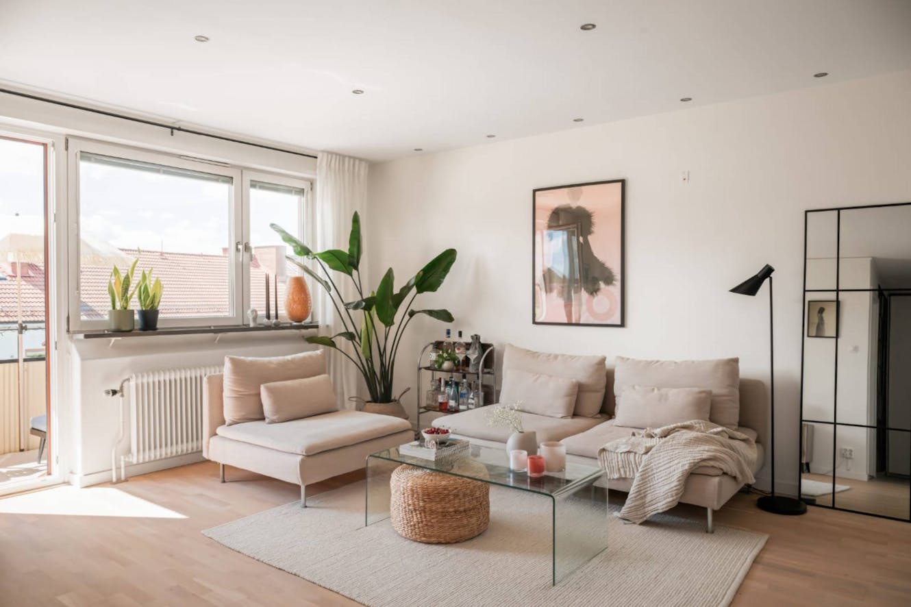 Living room with beige sectional sofa, glass coffee table with woven ottoman, large windows with natural light, potted plants, and modern floor lamp