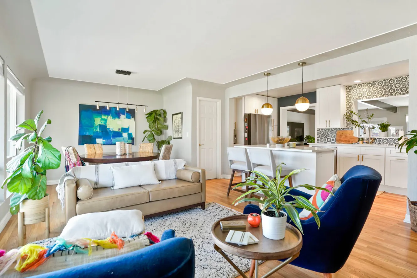 Living room with beige sofa, blue velvet chairs, round coffee table, hardwood floors, and open concept kitchen with white cabinets and pendant lights