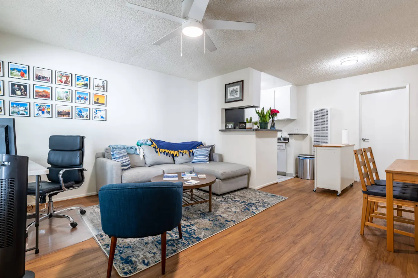 Living room with gray sectional sofa, blue accent chair, wood coffee table, ceiling fan, gallery wall with framed photos, and open kitchen area with white cabinets