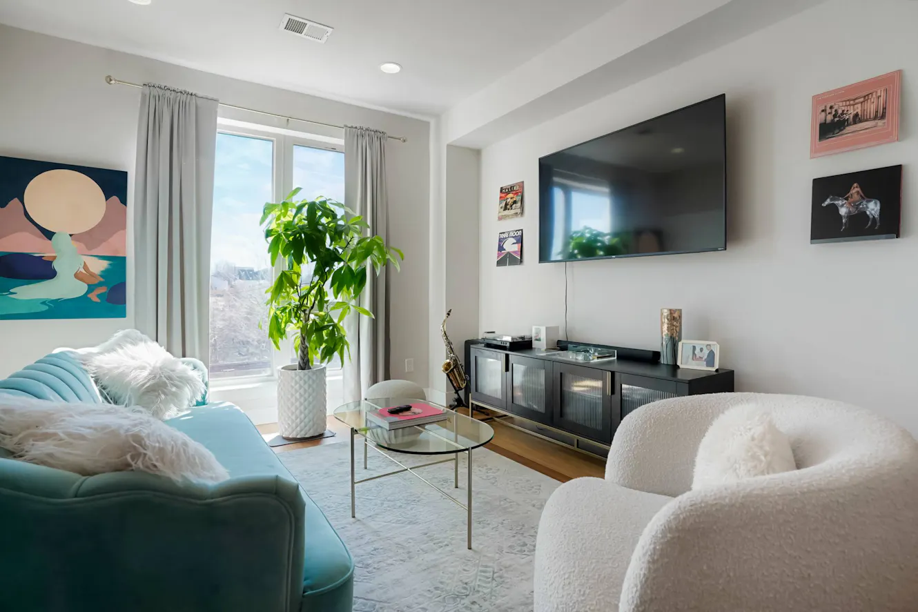 Living room with turquoise sofa, white armchair, wall-mounted TV, black media console, glass coffee table, large window with mountain view, and indoor plant