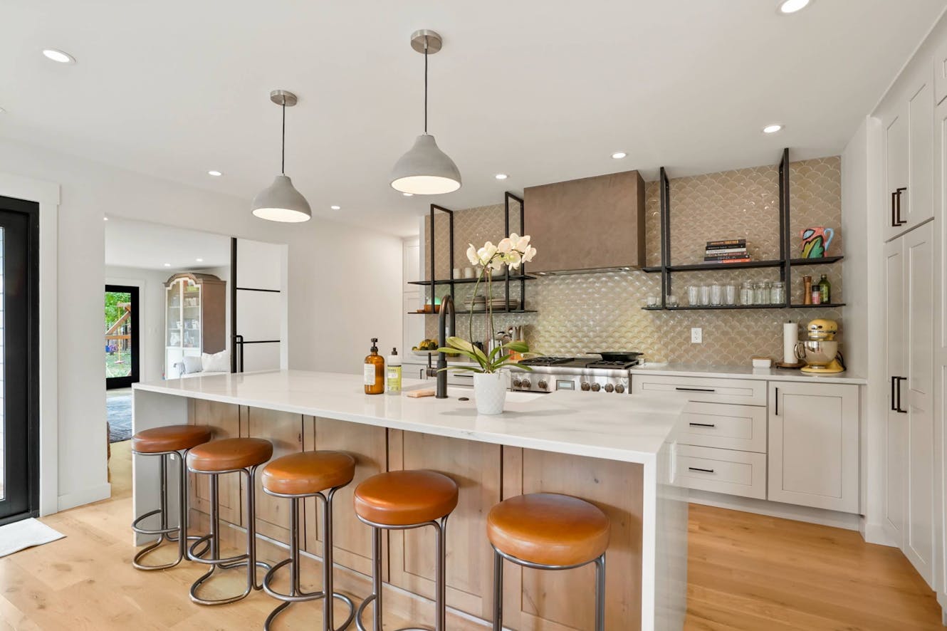 Kitchen with white quartz island, five leather bar stools, pendant lighting, gas range, open shelving, and geometric tile backsplash