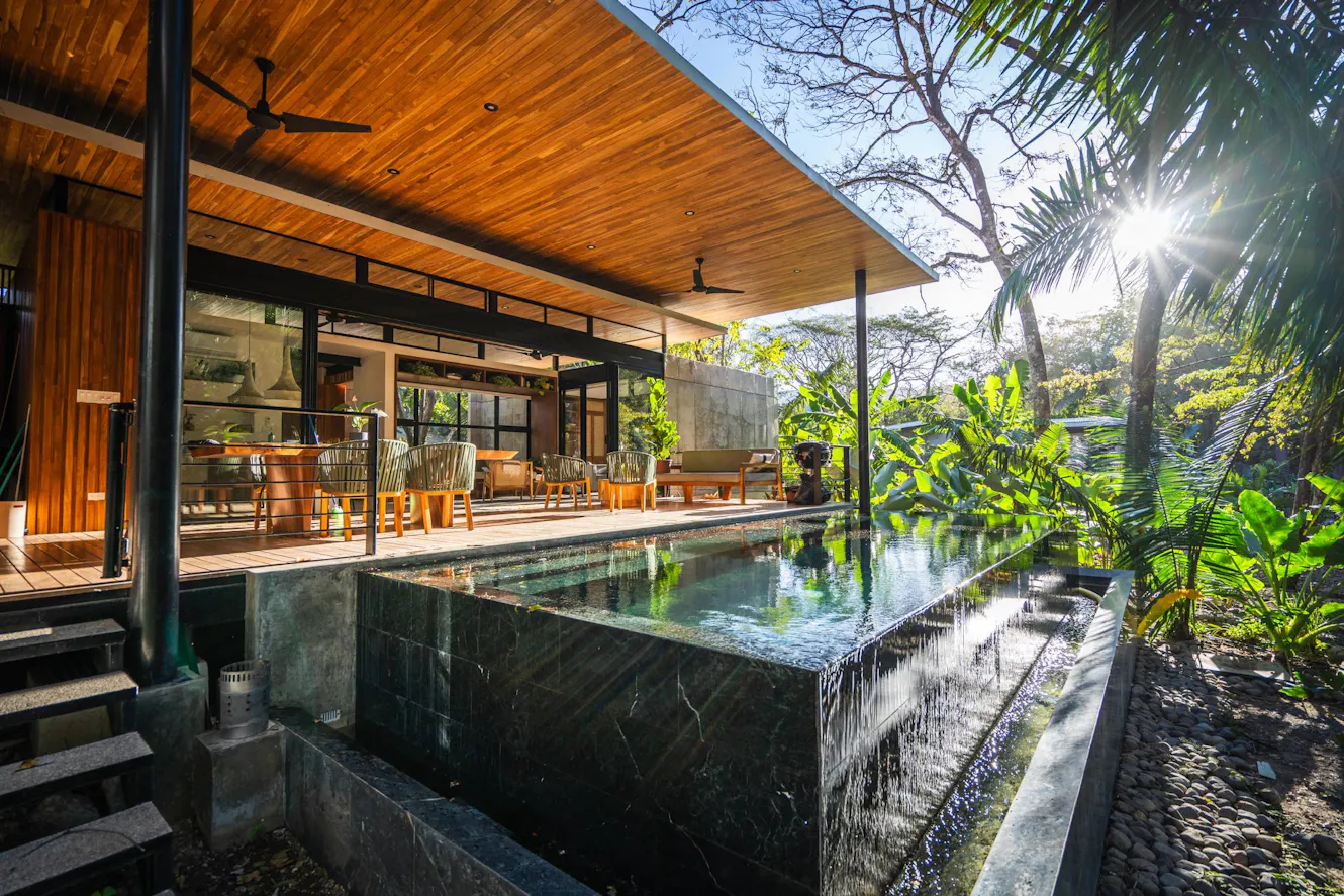 Modern pool with waterfall feature, wooden deck ceiling with ceiling fans, outdoor dining area, and tropical landscaping with palm trees
