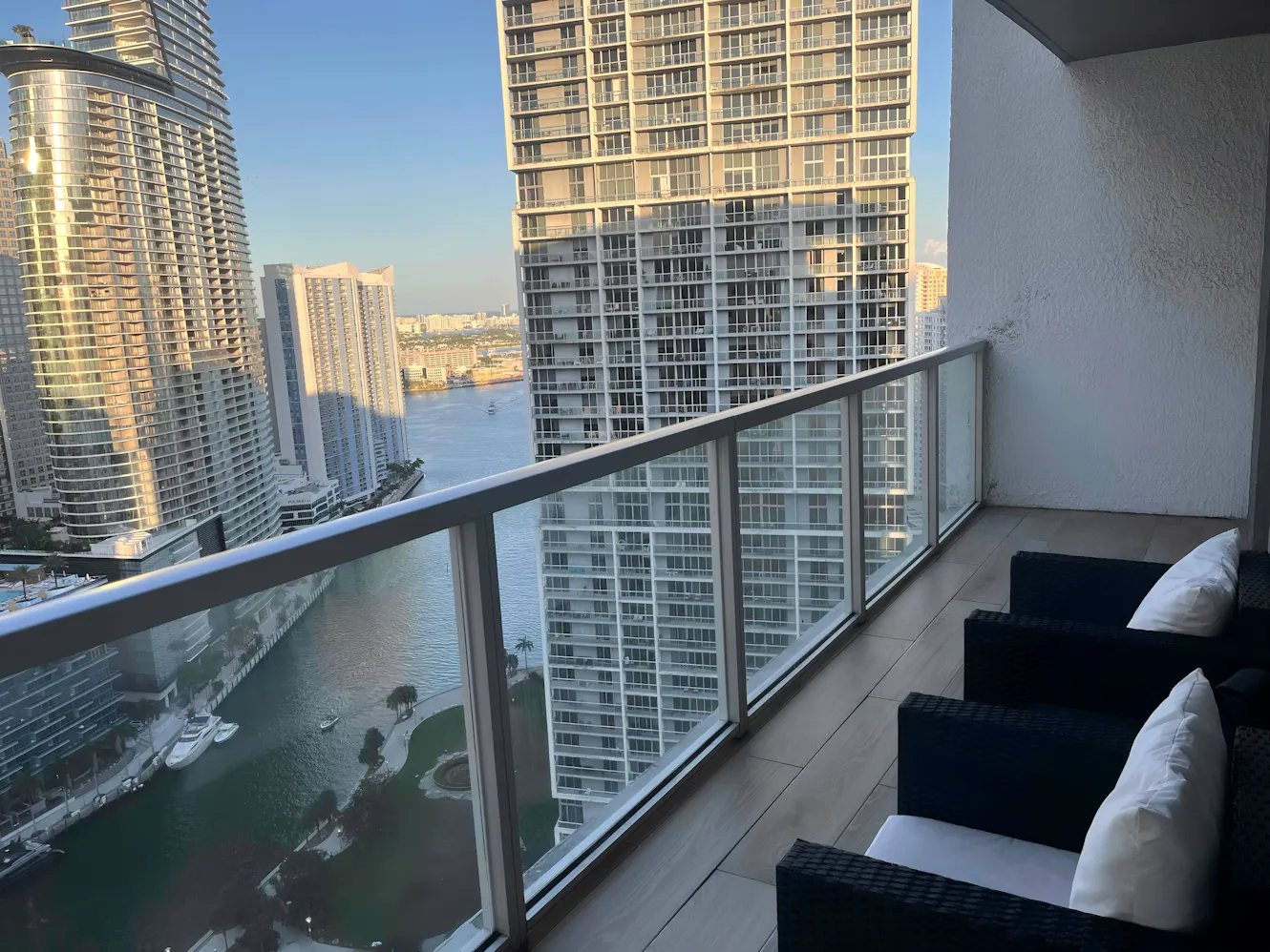 Balcony with glass railing, black wicker outdoor furniture with white cushions, and panoramic city and waterway views
