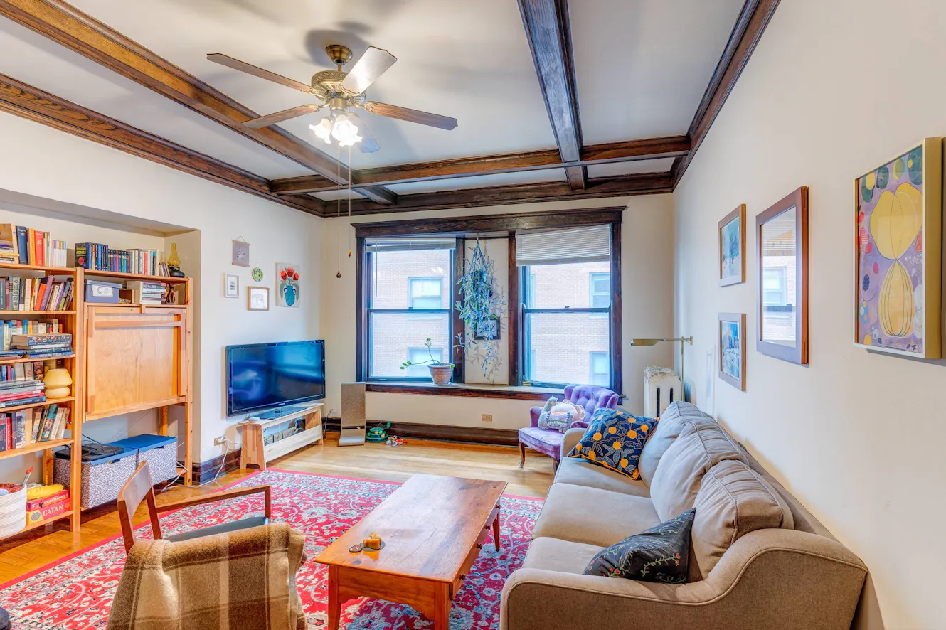 Living room with beige sofa, wooden coffee table, purple accent chair, bookshelf, wall-mounted TV, exposed wood beam ceiling, and large windows