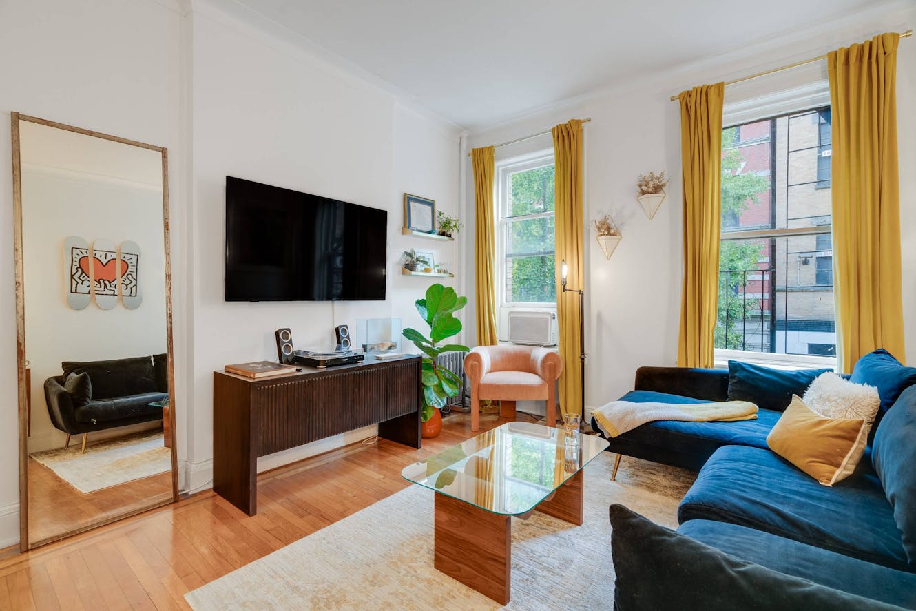 Living room with blue velvet sofa, glass coffee table, wall-mounted TV above media console, two windows with yellow curtains, and large floor mirror