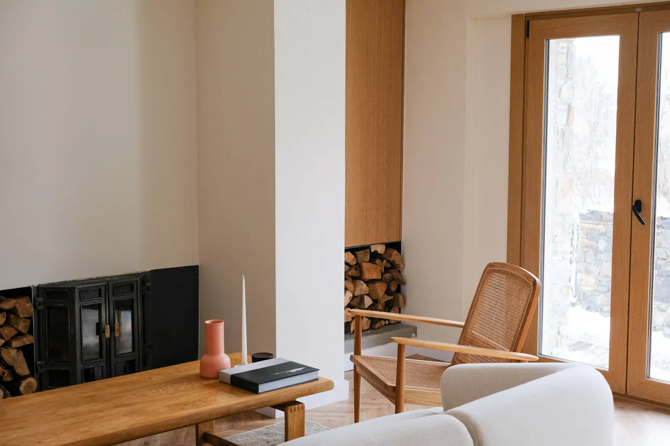Living room with wood-burning fireplace, wooden coffee table, woven accent chair, and glass door with winter view