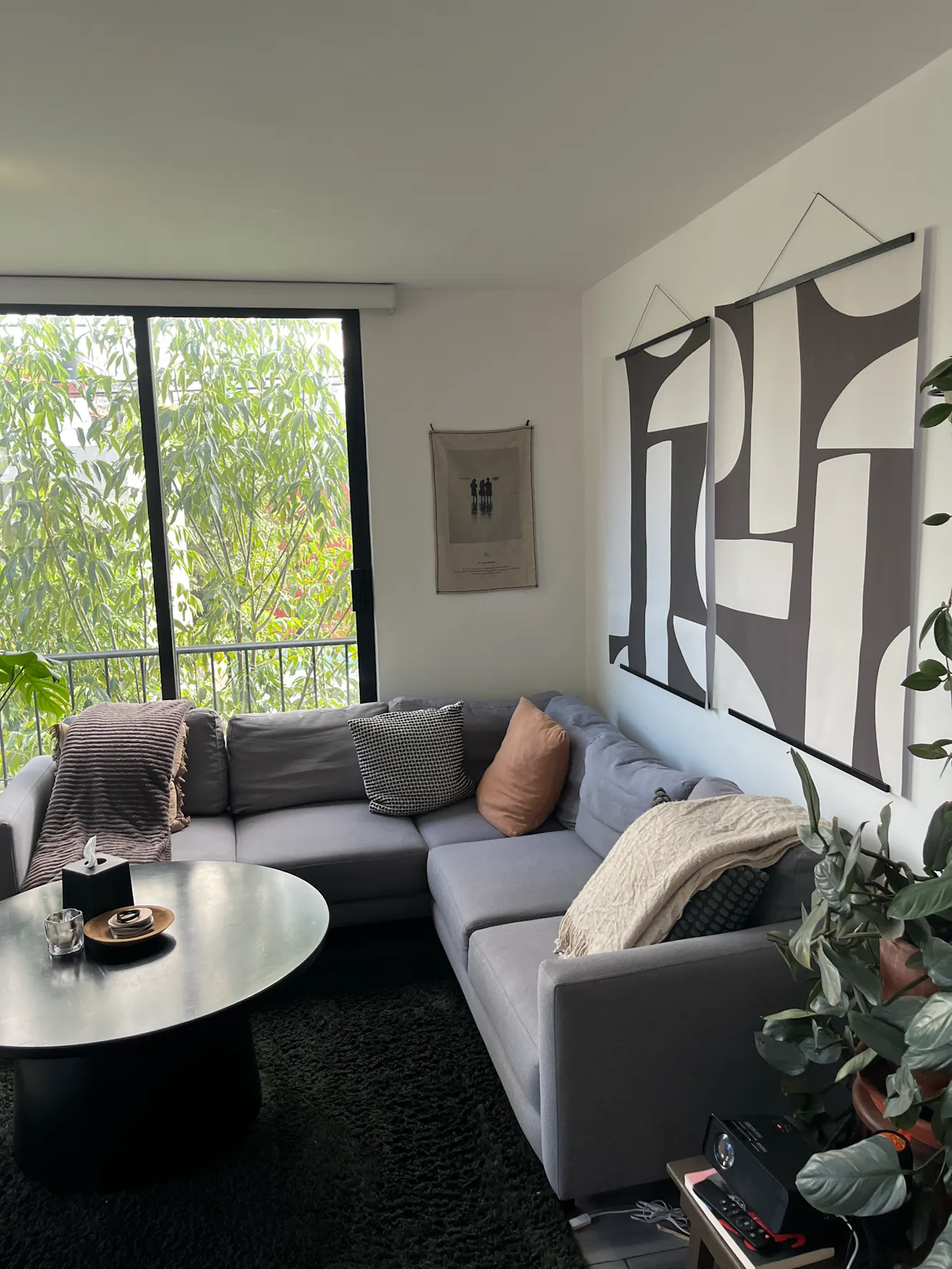 Living room with gray sectional sofa, round white coffee table, large window with tree view, modern wall art, and indoor plants