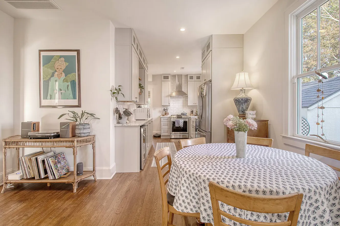 Dining room with round wooden table and chairs, large window with natural light, console table with record player, and view into white kitchen with stainless appliances