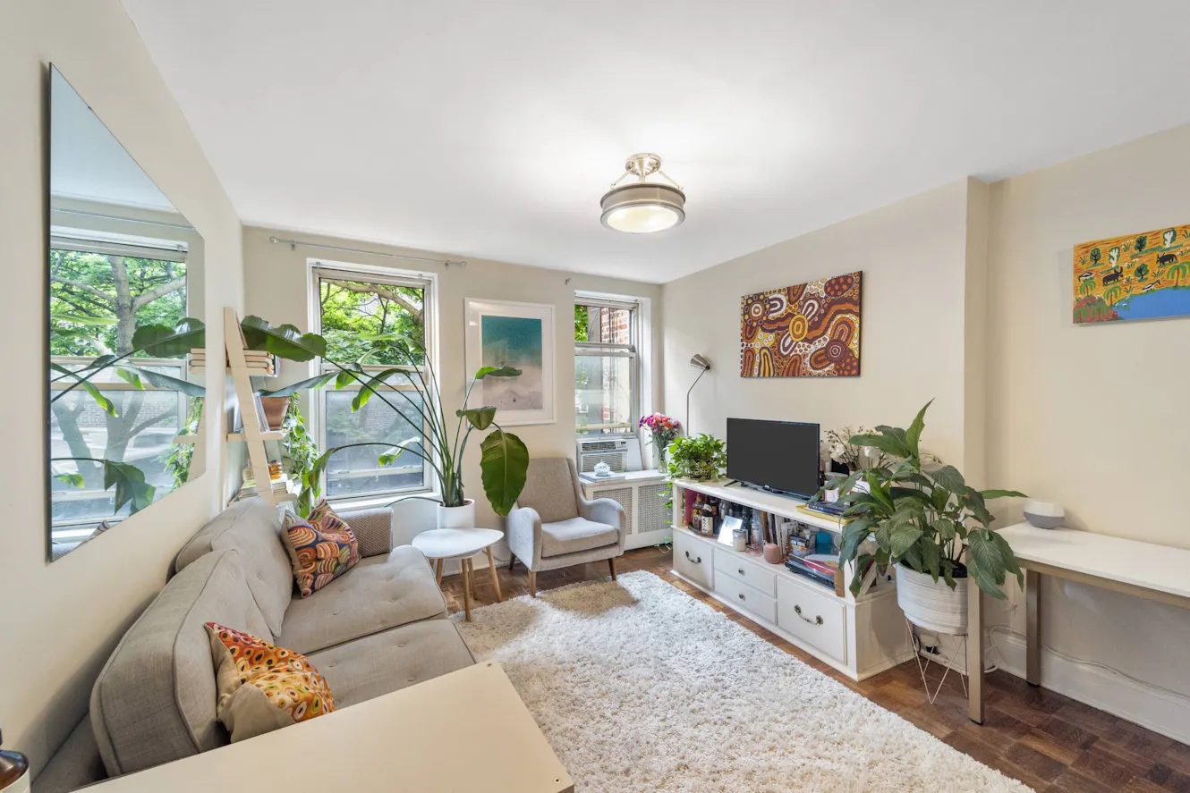 Living room with beige sectional sofa, white area rug, TV console, armchair, windows with natural light, indoor plants, and colorful wall art