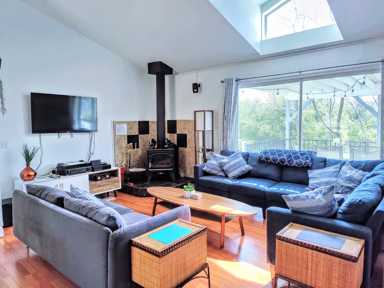 Living room with gray and black sectional sofas, wall-mounted TV, wood-burning stove, skylight, wooden coffee table, and large windows overlooking trees