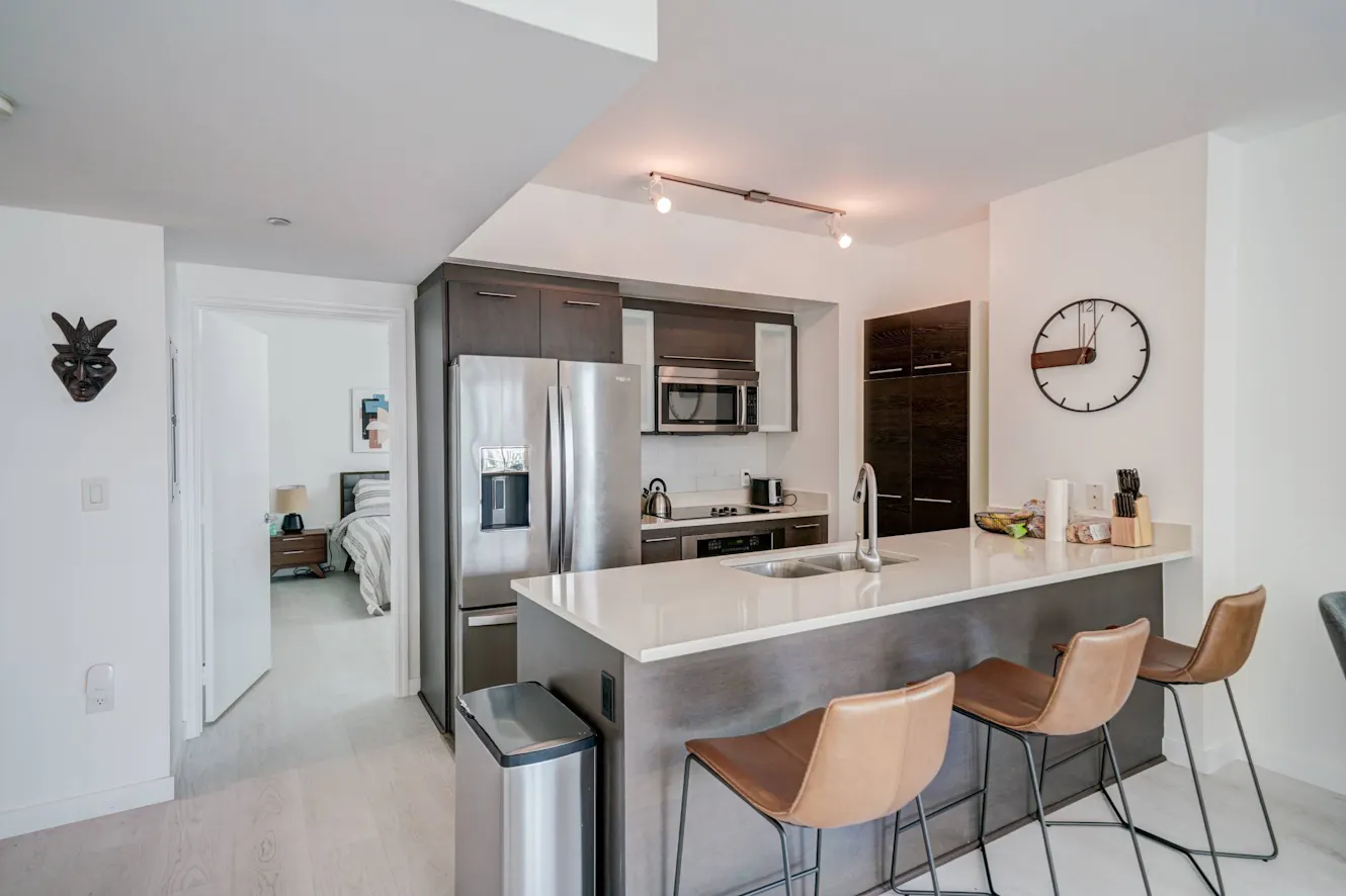 Kitchen with white quartz countertop island, brown bar stools, stainless steel appliances, dark wood cabinets, and track lighting