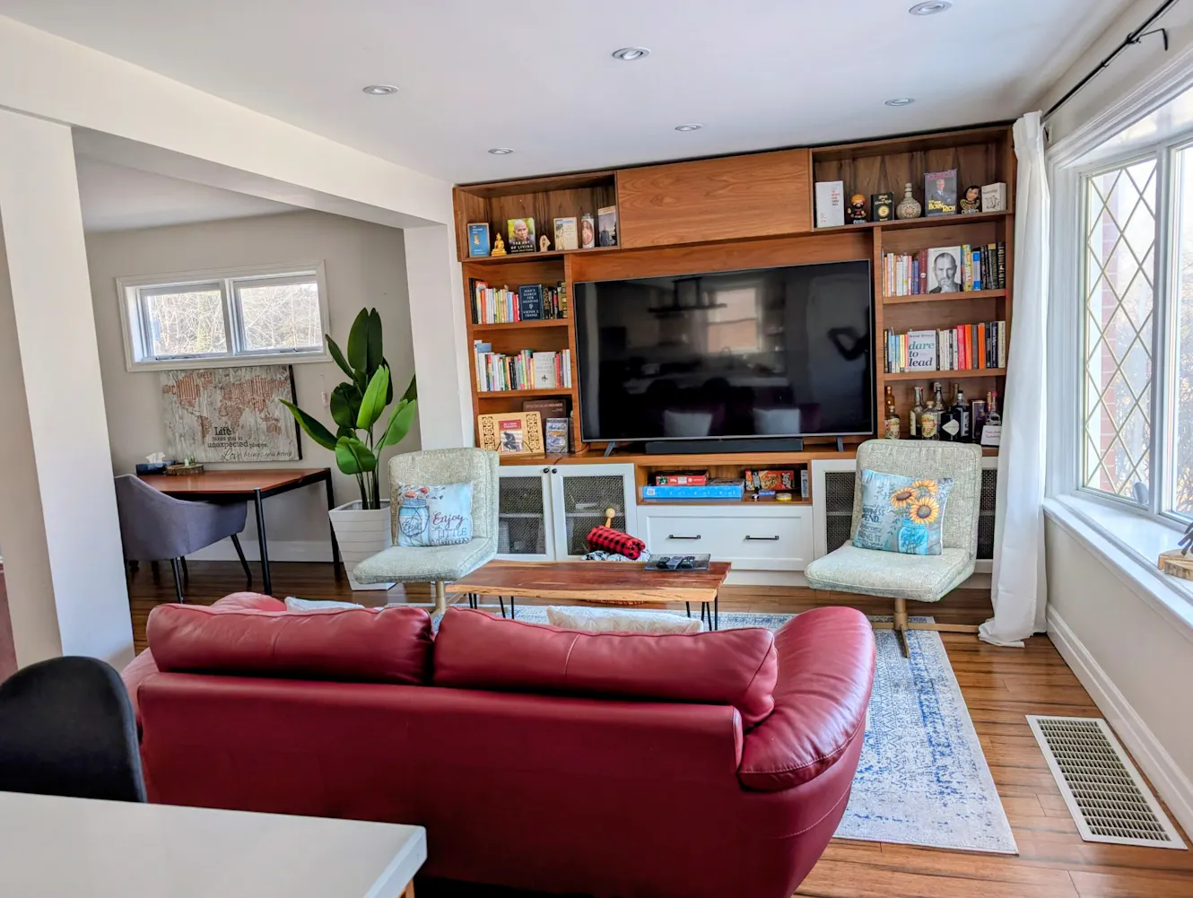 Living room with red leather sofa, wooden entertainment center with wall-mounted TV, two accent chairs, coffee table, hardwood floors, and large windows with natural light