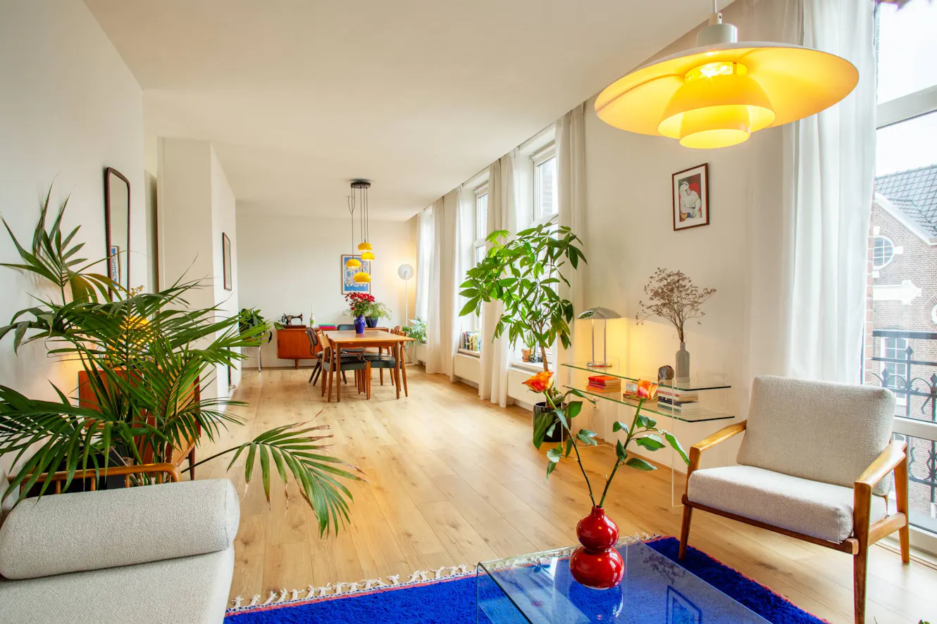 Living room with light wood floors, white armchairs, large yellow pendant light, floor-to-ceiling windows with white curtains, and indoor plants throughout