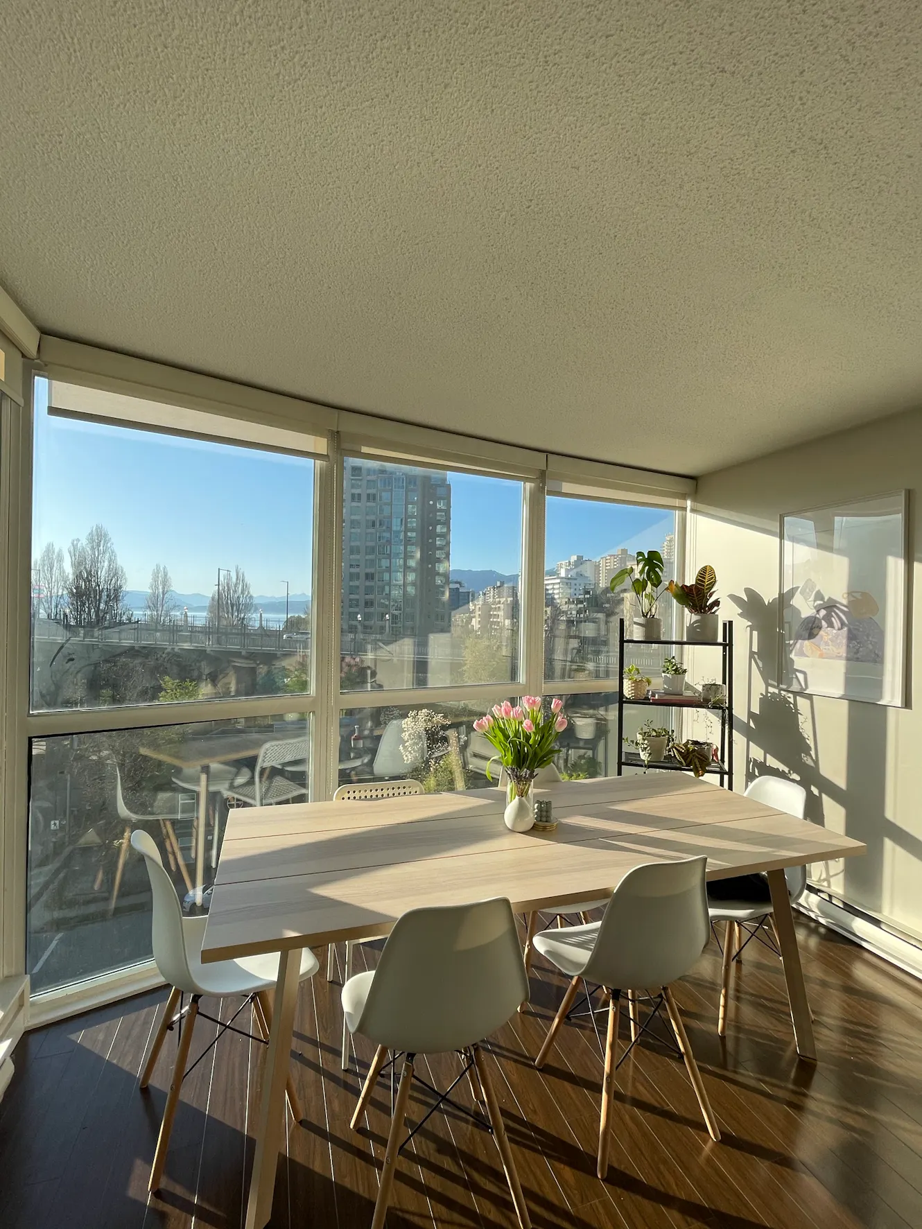Dining room with light wood table, white chairs, floor-to-ceiling windows with city and water views, plant shelf, and hardwood floors